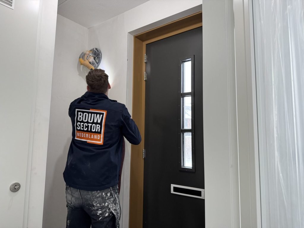 Vakman van Bouwsector Nederland in werkjas schuurt de wand boven een voordeur, de vloer is afgedekt