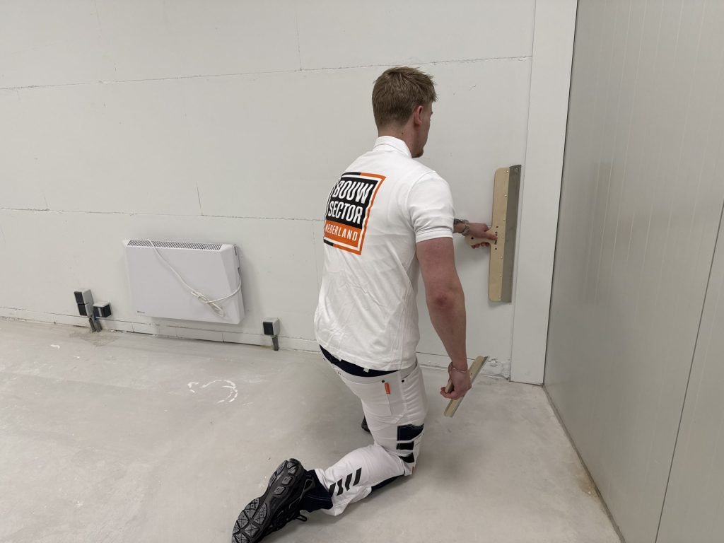 Stukadoor van Bouwsector Nederland werkt geknield het onderste deel van een kale wand af in een nieuwe woning